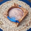 Ceniza y Mar: Una Pieza Artesanal que Captura la Esencia Mediterránea Para quienes disfrutan de un momento de pausa y reflexión, este accesorio eleva la experiencia a un plano superior. Nuestro cenicero cuadrado (12×12×4 cm) es una creación que denota alta distinción y aprecio por los objetos únicos. Fabricado completamente a mano, utiliza la robusta y aromática madera de Olivo por sus vetas singulares y su carácter inconfundible. Su principal atractivo reside en la armonía con la Resina Epoxi Azul, una combinación muy solicitada que recrea la profundidad y la luminosidad de las aguas costeras. El diseño es intencionalmente práctico y depurado, permitiendo que la belleza de los materiales y el acabado hablen por sí mismos. Este cenicero es la pieza de conversación ideal para cualquier ambiente, desde el interiorismo más selecto hasta una terraza cuidada. Es un complemento con gran personalidad para quienes valoran la durabilidad y la exquisita manufactura artesana, ofreciendo un soporte digno para el ritual del fumador.