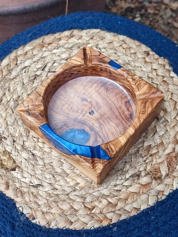 Ceniza y Mar: Una Pieza Artesanal que Captura la Esencia Mediterránea Para quienes disfrutan de un momento de pausa y reflexión, este accesorio eleva la experiencia a un plano superior. Nuestro cenicero cuadrado (12×12×4 cm) es una creación que denota alta distinción y aprecio por los objetos únicos. Fabricado completamente a mano, utiliza la robusta y aromática madera de Olivo por sus vetas singulares y su carácter inconfundible. Su principal atractivo reside en la armonía con la Resina Epoxi Azul, una combinación muy solicitada que recrea la profundidad y la luminosidad de las aguas costeras. El diseño es intencionalmente práctico y depurado, permitiendo que la belleza de los materiales y el acabado hablen por sí mismos. Este cenicero es la pieza de conversación ideal para cualquier ambiente, desde el interiorismo más selecto hasta una terraza cuidada. Es un complemento con gran personalidad para quienes valoran la durabilidad y la exquisita manufactura artesana, ofreciendo un soporte digno para el ritual del fumador.